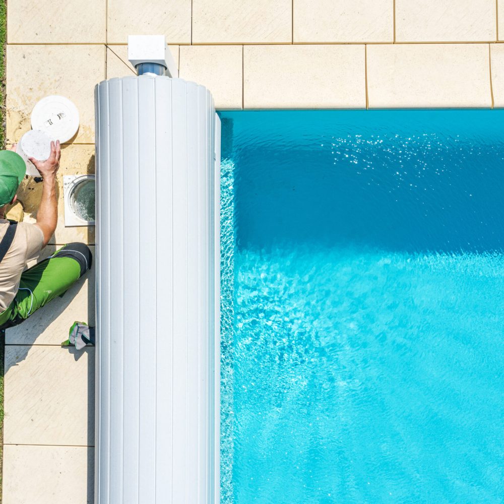 A professional is checking the pool water quality beside a sparkling blue pool in a residential backyard. The grass is lush and green, indicating a warm, sunny day.